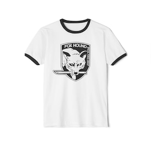 Puede incluir: Camiseta blanca con ribete negro. La parte delantera muestra un gráfico en blanco y negro de una cabeza de zorro sosteniendo un cuchillo, con el texto "FOX HOUND" y "SPECIAL FORCE GROUP".
