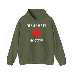 May include: An olive green hoodie with a front pocket. The front displays the text "M*A*S*H" above a red cross and the number "4077TH" in white. The hoodie has a drawstring and a relaxed fit, typical of a sweatshirt.