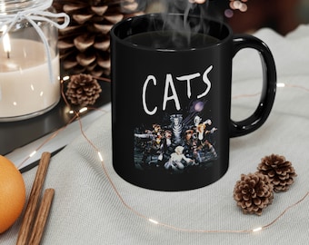 May include: Black coffee mug with a white "CATS" graphic and a photo of the cast of the musical Cats. The mug is filled with coffee and steam is rising from the mug.