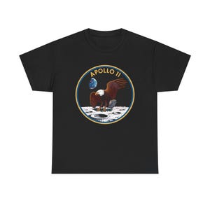Könnte beinhalten: Ein schwarzes Kurzarm-T-Shirt mit einem kreisförmigen Aufdruck auf der Brust. Das Motiv zeigt einen Weißkopfseeadler, der auf der Mondoberfläche landet, während die Erde am dunklen Himmel zu sehen ist. Der Text APOLLO 11 steht in goldenen Buchstaben oben.