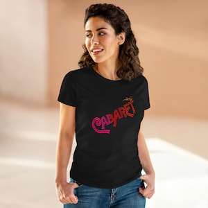 Può includere: T-shirt nero con la scritta "CABARET" in un design a pois rosa e rosso. La maglietta ha maniche corte e girocollo. La modella indossa jeans blu.