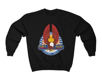 Pode incluir: Camisola preta com um design gráfico colorido. Apresenta uma águia com as asas abertas, uma estrela e o texto "FALL GUY STUNTMAN ASSOC" num círculo. A águia está sobre um fundo vermelho, branco e azul.