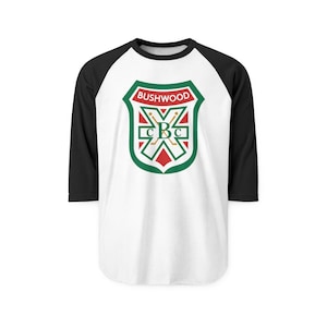 May include: White baseball tee with black sleeves. The shirt features a green, red, and gold crest with the word "BUSHWOOD" at the top and the letters "CBC" in the center. The shirt has a crew neck and three-quarter length sleeves.