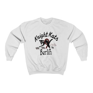 May include: White sweatshirt with a black and red graphic of a cat with the text "Knight Kats 9 Lives Berlin".