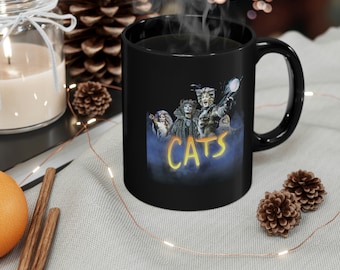May include: Black coffee mug with a graphic of characters from the musical Cats. The mug has the word "CATS" in yellow letters on the front.