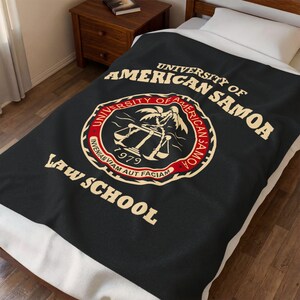 Puede incluir: Una manta de forro polar negra con el logotipo de la Facultad de Derecho de la Universidad de Samoa Americana en crema y rojo. El emblema circular muestra una palmera, una balanza de justicia, el año 1979 y la frase Inveniam Viam Aut Faciam.