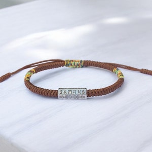 May include: A brown braided bracelet with a silver charm featuring Tibetan script. The bracelet has a multi-colored thread woven into the brown cord.