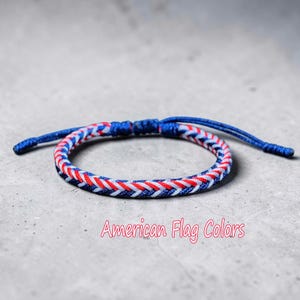 May include: A woven bracelet in red, white, and blue, reminiscent of the American flag. The bracelet features a braided pattern and a blue adjustable fastening. The text "American Flag Colors" is visible.