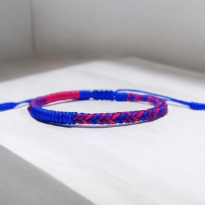May include: A blue, pink, and purple braided friendship bracelet. The bracelet is made with a woven knot pattern.