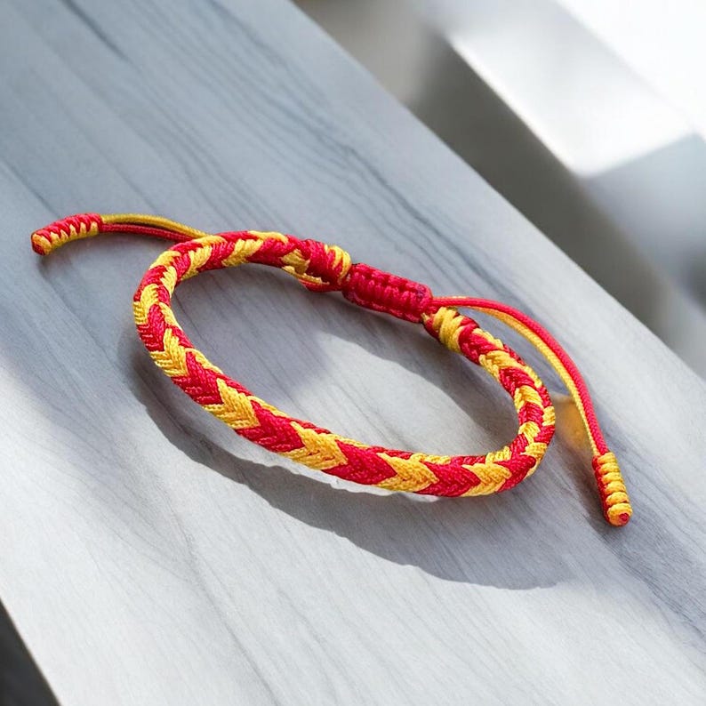 May include: A red and yellow braided friendship bracelet. The bracelet is made of two strands of cord, one red and one yellow, that are braided together in a chevron pattern.