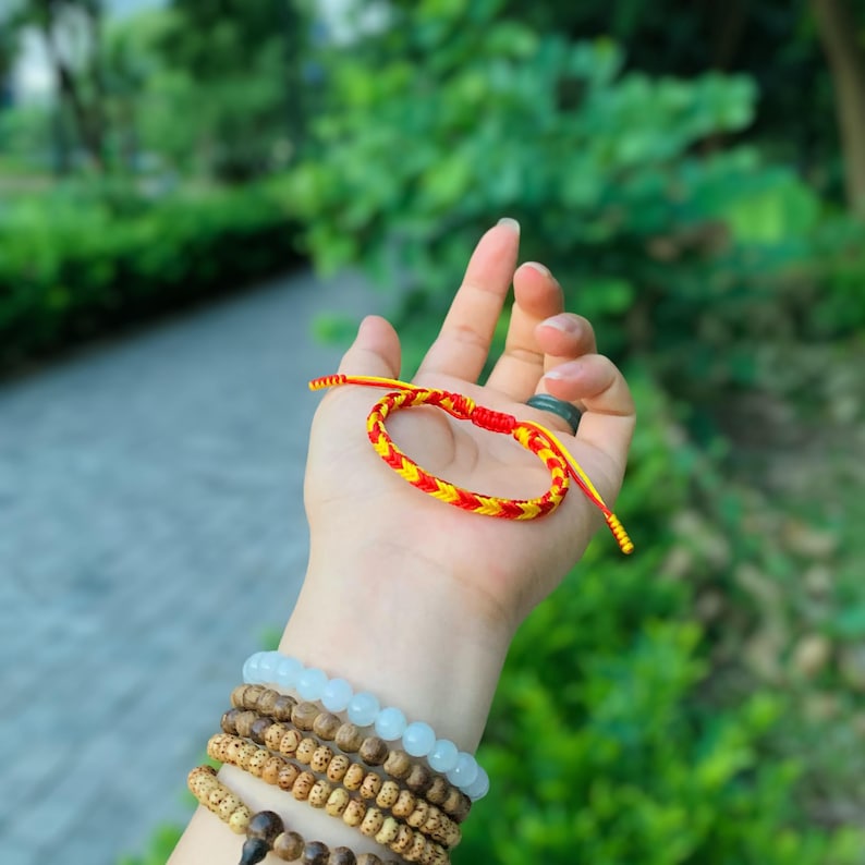 May include: A red and yellow braided string bracelet with a knot closure. The bracelet is held in a hand with other bracelets on the wrist.
