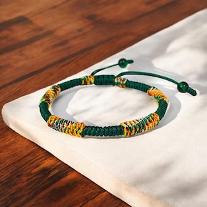 May include: A green woven bracelet with colorful accents of yellow, orange, and white. The bracelet features a sliding knot closure with green beads. The bracelet is displayed on a white surface with a wooden background.
