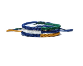 May include: Three braided bracelets in blue, white, green, and gold colors. The bracelets are stacked on top of each other.