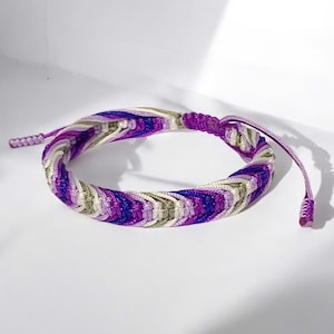 May include: Braided bracelet in shades of purple, white, and silver. The bracelet features an adjustable sliding knot closure. The woven design creates a textured appearance. The bracelet is a fashion accessory.