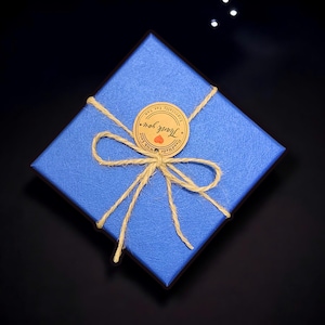 Op de afbeelding: Een vierkante, blauwe cadeauverpakking met textuur, vastgebonden met touw. Een ronde sticker met de woorden "Thank you" en een hart is gecentreerd op de doos. De doos staat op een zwart oppervlak.