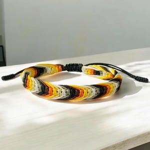 Pode incluir: Pulseira trançada com uma mistura de fios amarelos, laranja, pretos e brancos. A pulseira tem um design tecido e cordões pretos ajustáveis para fechar. A pulseira é exibida em uma superfície branca, mostrando suas cores vibrantes e seu artesanato intrincado.