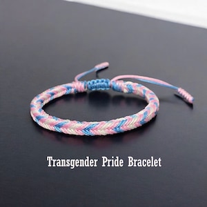 May include: A braided friendship bracelet in light blue, pink, and white colors. The bracelet is tied in a knot at the end. The text "Transgender Pride Bracelet" is written below the bracelet.