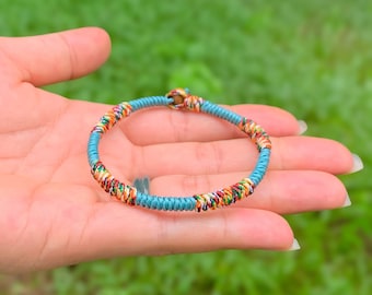 May include: A handmade bracelet with a turquoise blue cord and colorful woven accents. The bracelet features a vibrant mix of orange, yellow, red, and green threads, creating a unique and eye-catching design. The bracelet is held in a hand against a blurred green background.