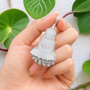 May include: A white jade pendant carved in the shape of a seated Buddha figure. The pendant is encased in a silver-toned metal frame accented with small, clear, sparkling stones along the edges and the bail at the top of the piece.