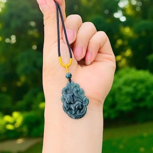 Op de afbeelding: Een donkergroene hangerketting met een gedetailleerd gesneden ontwerp. De hanger hangt aan een zwart koord met een goudkleurige sluiting. De hanger heeft een gesneden dierfiguur.