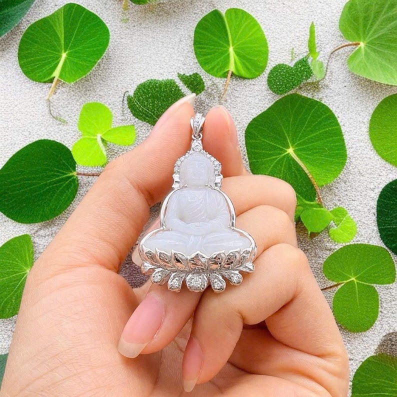 May include: A white jade Buddha pendant set in a silver-toned metal frame with small clear stones. The Buddha figure is depicted in a seated meditation pose on a lotus base, with a decorative bail at the top for a necklace chain attachment.
