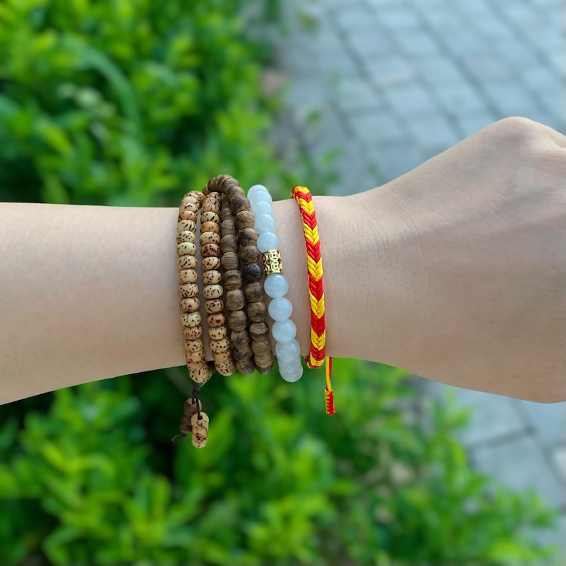 May include: A set of five bracelets on a wrist. The bracelets are made of wood beads, white beads, and red and yellow string.