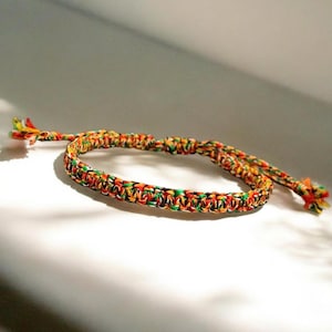 Könnte beinhalten: Ein handgefertigtes Armband mit einem gewebten Design in einer lebendigen Mischung aus Farben: Rot, Gelb, Grün und Orange. Das Armband ist verstellbar mit geflochtenen Enden und einem Schiebeknotenverschluss.