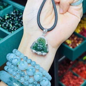 Peut inclure: Ensemble de bijoux en jade comprenant un pendentif Bouddha vert sur un cordon noir, une bague et plusieurs bracelets. Les bracelets présentent des perles rondes et cylindriques dans des tons de bleu et de vert.