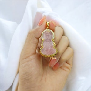 May include: A gold pendant featuring a pink crystal carving of a seated figure with a halo of clear crystals. The pendant is held in a hand with pink nail polish.