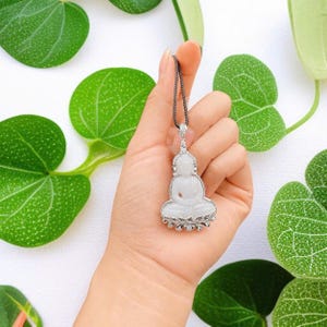 May include: A white jade Buddha pendant set in a silver frame with small clear stones, suspended from a thin dark chain. The pendant features a seated Buddha figure on a lotus base, with a decorative silver bail at the top connecting to the chain.