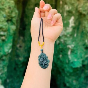 Op de afbeelding: Een donkergroene jade hangerketting met een gesneden ontwerp, hangend aan een zwart koord met een goudkleurige sluiting. De hanger wordt tegen een vage groene achtergrond gehouden.