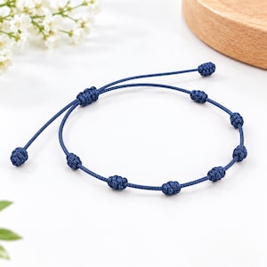 Puede incluir: Una pulsera de cordón azul oscuro que presenta una serie de nudos decorativos espaciados uniformemente a lo largo de la banda. La pulsera incluye un mecanismo de cierre deslizante ajustable con dos extremos sueltos, cada uno con un nudo.