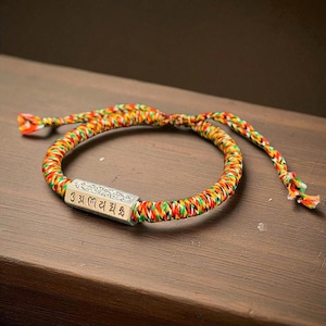 May include: A colorful braided bracelet with a silver rectangular charm. The bracelet is made of red, yellow, green, and orange threads. The charm has engraved text. The bracelet is on a brown wooden surface.
