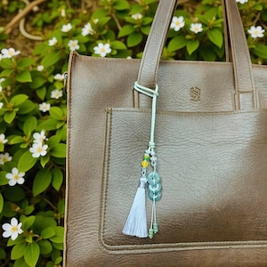 Puede incluir: Un bolso tote de cuero marrón con un adorno decorativo. El adorno presenta una borla azul claro, cuentas verdes y tres piedras de jade circulares. El bolso tiene una superficie texturizada y un bolsillo frontal.