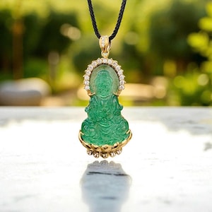 Può includere: Un ciondolo d'oro con una figura di Buddha verde scolpita. Il ciondolo presenta un'aureola di piccole pietre preziose trasparenti e una montatura in oro. È appeso a un cordino nero, che unisce spiritualità ed eleganza.