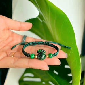 Könnte beinhalten: Ein grünes Jade-Armband mit einem grünen Jade-Anhänger in Form einer chinesischen Glücksmünze. Das Armband besteht aus grüner Schnur und hat goldene Perlen.