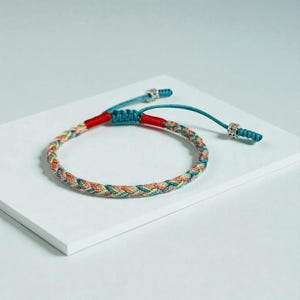 May include: A braided bracelet with a colorful pattern of orange, yellow, green, and blue threads. The bracelet has red accents and turquoise adjustable cords with silver-colored beads. The bracelet is displayed on a white surface.