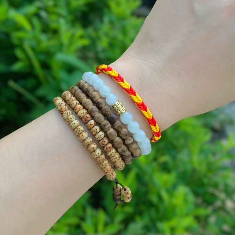 May include: A stack of bracelets on a wrist. The bracelets are made of wood beads, white beads, and a red and yellow braided string.