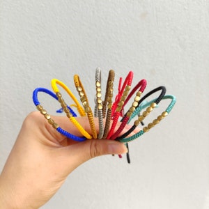 May include: A collection of colorful string bracelets with gold beads. The bracelets are made of different colors, including red, blue, yellow, green, and brown.