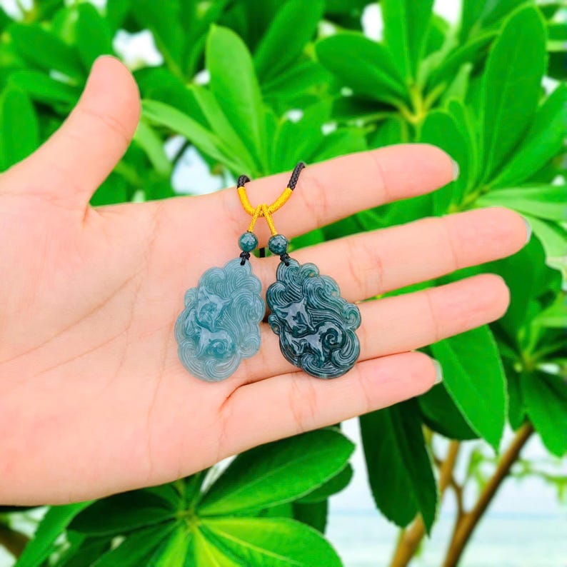 Op de afbeelding: Twee jade hangers met ingewikkelde gesneden ontwerpen, &eacute;&eacute;n lichtblauw en de andere donkergroen, hangen aan gele en zwarte koorden. De hangers worden getoond tegen een achtergrond van groen gebladerte, wat hun gedetailleerde vakmanschap benadrukt.