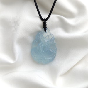 May include: A light blue carved jade pendant in the shape of a flower, hanging from a black cord necklace.