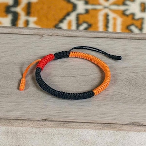 Può includere: Braccialetto fatto a mano con un design intrecciato. Il braccialetto presenta sezioni nere, arancioni e rosse. Il cordino regolabile è nero e arancione. Il braccialetto è esposto su una superficie di legno chiaro.