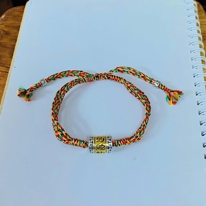 May include: A braided bracelet with a central cylindrical bead. The bracelet is made of colorful threads in red, yellow, green, and orange. The bead is gold-colored with etched symbols. The bracelet is on a white surface.