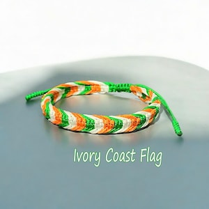 May include: Braided bracelet in the colours of the Ivory Coast flag: green, white, and orange. The bracelet is made of woven threads and has adjustable green cords. The text "Ivory Coast Flag" is displayed below the bracelet.