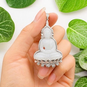 May include: A white jade Buddha pendant set in a silver-toned metal frame. The figure sits in a lotus position on a base adorned with small, clear, sparkling stones. The frame features a decorative bail at the top, also accented with clear stones.