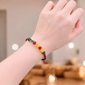 May include: A red, yellow, and green braided bracelet with decorative knots. The bracelet features a series of colorful knots in yellow, red, and green, with a dark red cord. The bracelet is worn on a wrist.