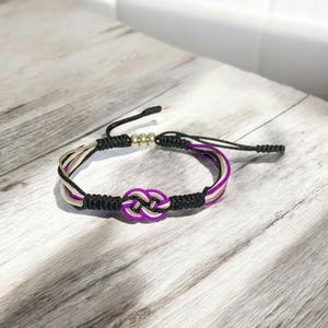 May include: A handmade bracelet featuring a central knot design in purple, black, and gold. The bracelet has adjustable black cords and metallic beads. The design is set against a light-colored wooden surface.