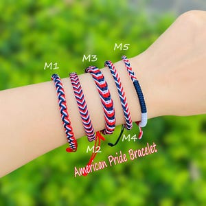Puede incluir: Cinco pulseras trenzadas en rojo, blanco y azul, con el texto "American Pride Bracelet". Las pulseras presentan diferentes patrones, con los colores dispuestos en rayas y chevrones. Las pulseras están en una muñeca.