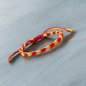 May include: A red and yellow braided friendship bracelet. The bracelet is made of two strands of cord that are woven together in a simple pattern.