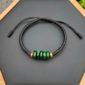 May include: A black braided cord bracelet featuring five green beads and two gold-colored beads. The bracelet is adjustable with sliding knots. The beads are a vibrant green color.